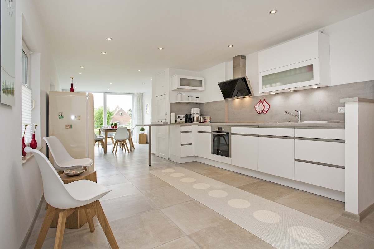 A modern kitchen features sleek white cabinetry and a spacious countertop. Natural light floods the area through large windows. A dining table with chairs is visible in the background. A decorative rug with circular patterns lies on the tiled floor, enhancing the minimalist design.