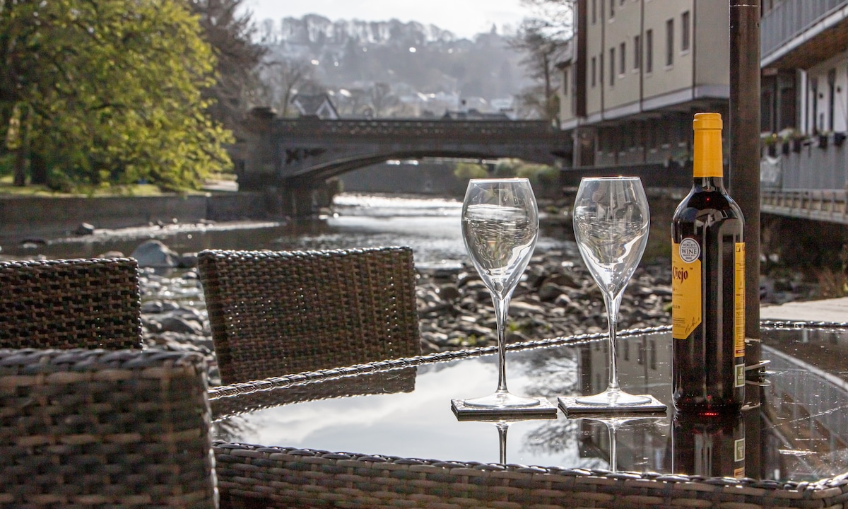 A table set for outdoor dining features two glasses and a bottle on a reflective surface. In the background, the serene River Greta flows past, framed by greenery and distant buildings, with a bridge crossing the water.