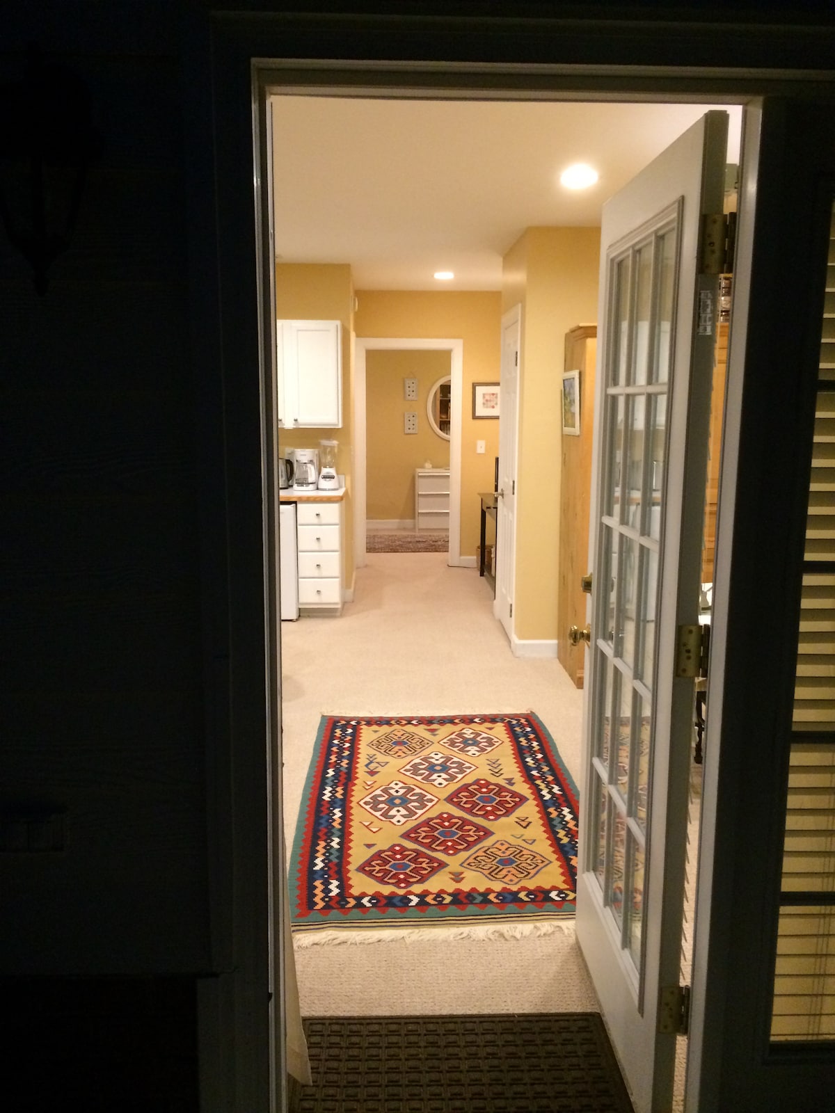 A view from the entrance reveals a welcoming interior. Soft yellow walls surround a pathway leading to a well-appointed kitchen. A colorful area rug lays across the entryway, while artistic decor and practical furnishings complement the space.