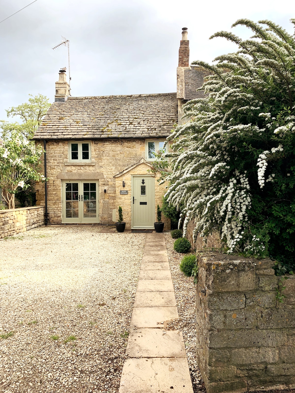 A charming stone cottage is framed by lush greenery and flowering shrubs, creating a welcoming entrance. The well-defined pathway leads to a stylish front door, while a gravelled area provides off-street parking. The overall aesthetic combines rustic character with well-maintained landscaping.