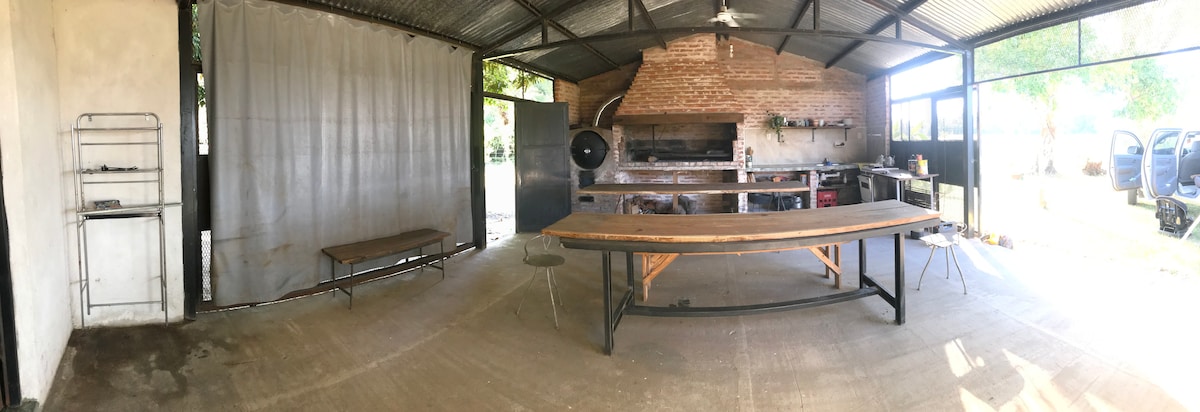 A spacious semicovered area features a large table and a grill setup built into a rustic brick wall. Additional cooking amenities are present, including a wood oven and gas stove. Ample natural light enters through large openings, enhancing the inviting space.