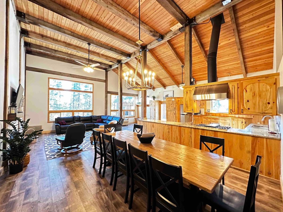 An open-concept dining and kitchen area is showcased, featuring a large wooden dining table with black chairs. The kitchen features wooden cabinetry, a central island, and a range hood. Ceiling beams and large windows allow natural light to illuminate the space, creating a warm atmosphere.