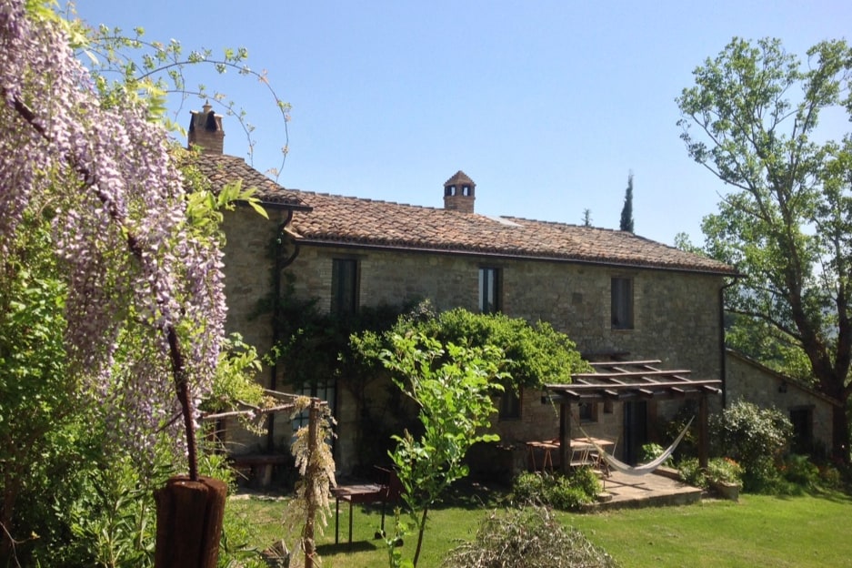 The exterior of an antique stone farmhouse is shown, surrounded by lush greenery. A pergola is visible in the garden, complemented by a hammock. Trees and climbing plants frame the building, which features a rustic tiled roof and several windows reflecting natural light.