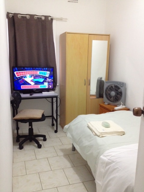 The compact room features a spacious bed covered with a white blanket. A desk with a chair and a computer monitor is positioned against one wall. A wardrobe and a small wooden nightstand are visible, along with a fan providing air circulation. Natural light filters in through a nearby window.