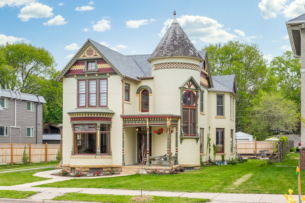 The exterior of a Victorian-style house showcases intricate architectural details, including a turret and decorative trim. Well-maintained gardens with flower beds adorn the front yard, while a paved pathway leads to the welcoming entrance. Lush greenery surrounds the property, enhancing its appeal.