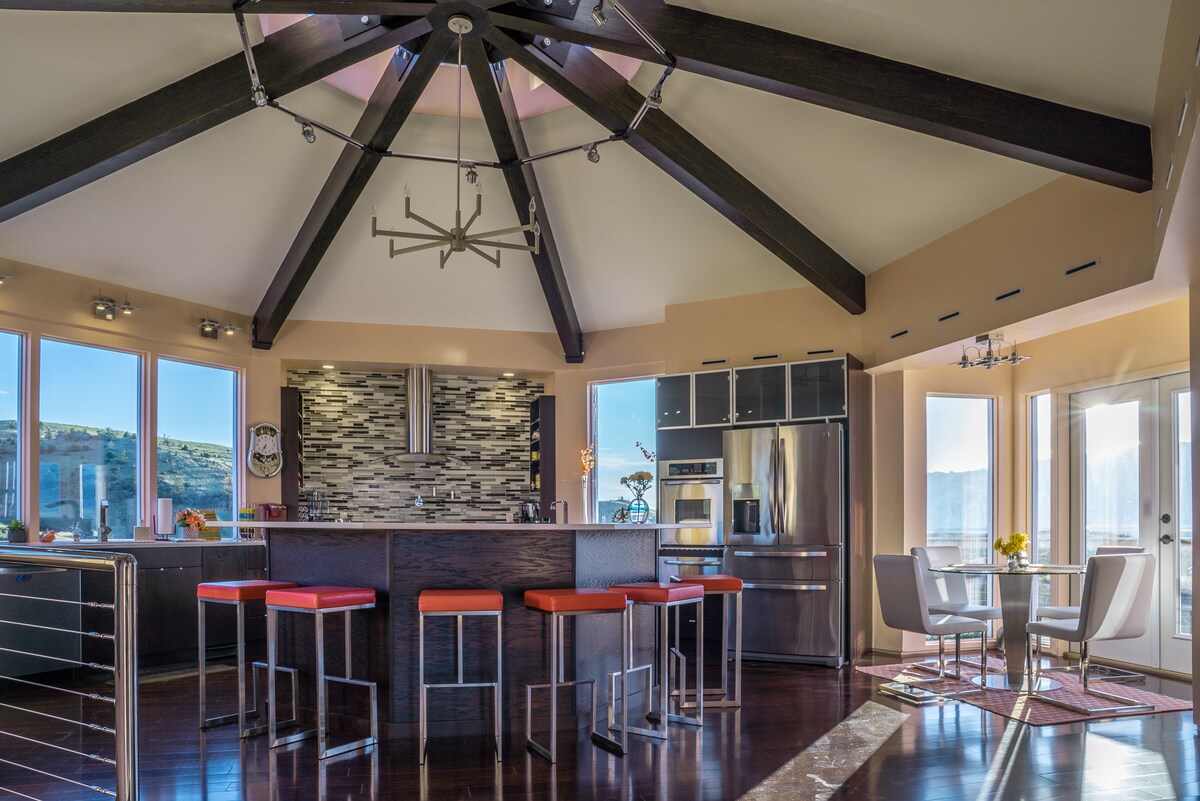 The kitchen area features a modern design with a central island surrounded by four tall stools. A striking ceiling showcases wooden beams, while large windows provide ample natural light and scenic mountain views. Stainless steel appliances and stylish cabinetry complete the space.