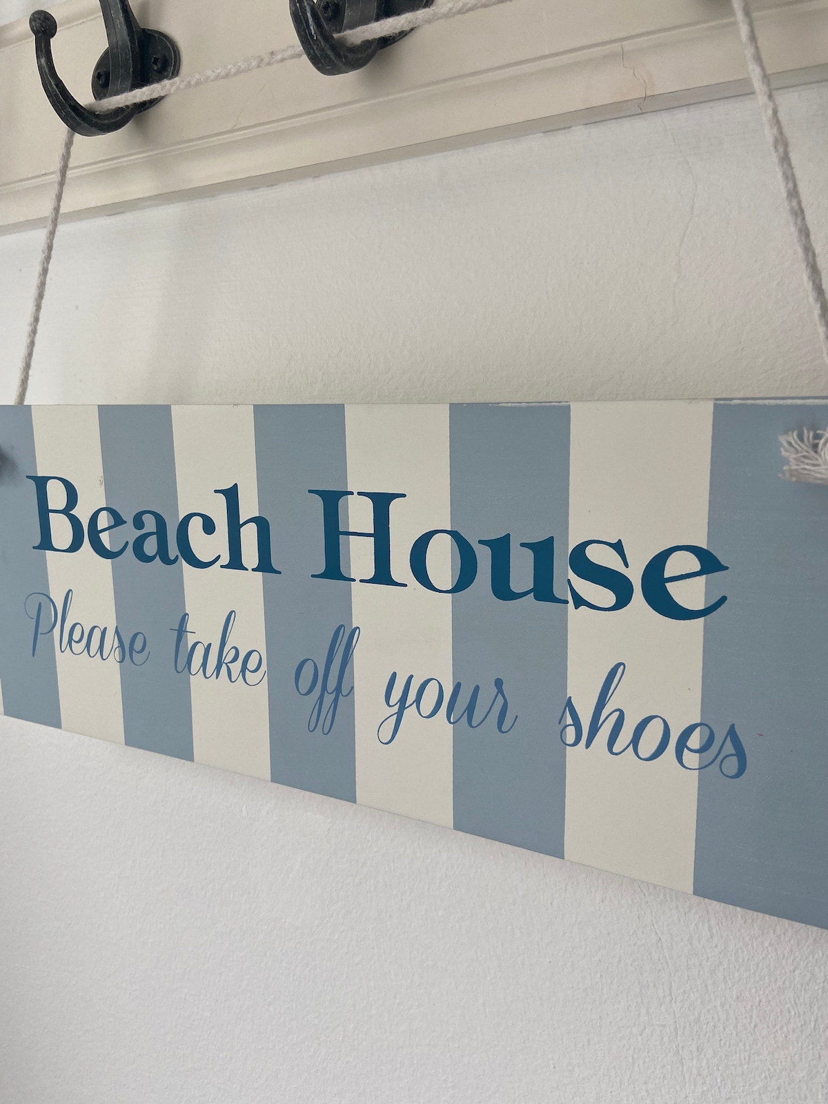 A light blue and white striped sign reads 'Beach House' and 'Please take off your shoes' in an inviting font. The sign is hung by twine from a simple hook, providing a welcoming atmosphere at the entrance.