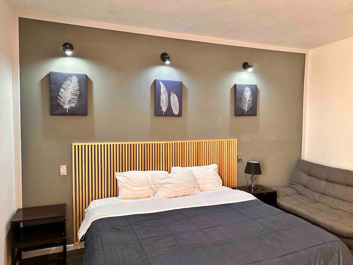 A spacious sleeping area features a king size bed with a premium mattress and soft linens. Above the bed, three framed leaf prints are illuminated by sconce lights. A gray sofa occupies the corner, providing additional comfort.