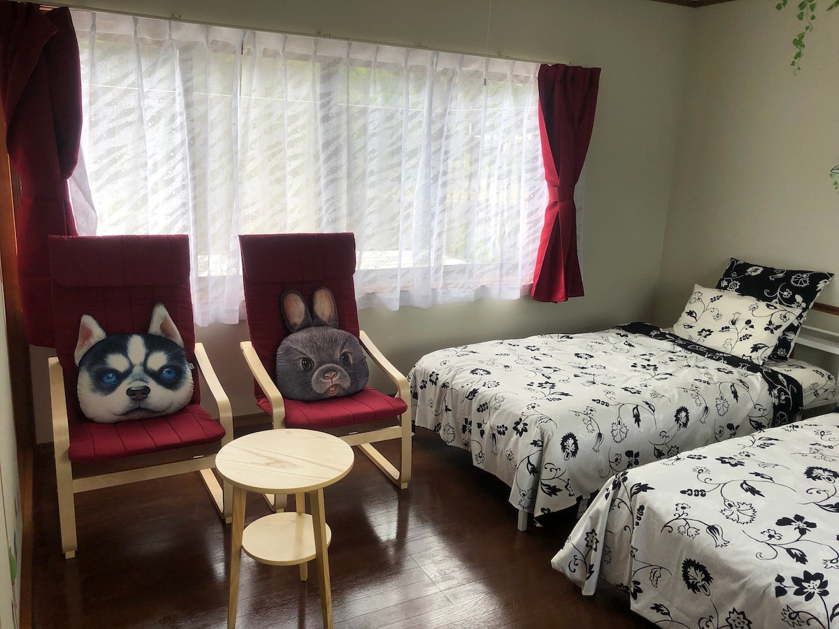 A bright room features two single beds dressed in floral patterned bedding. Two red armchairs, adorned with animal-themed cushions, face a small wooden table. Natural light filters through sheer curtains, highlighting the simple decor.
