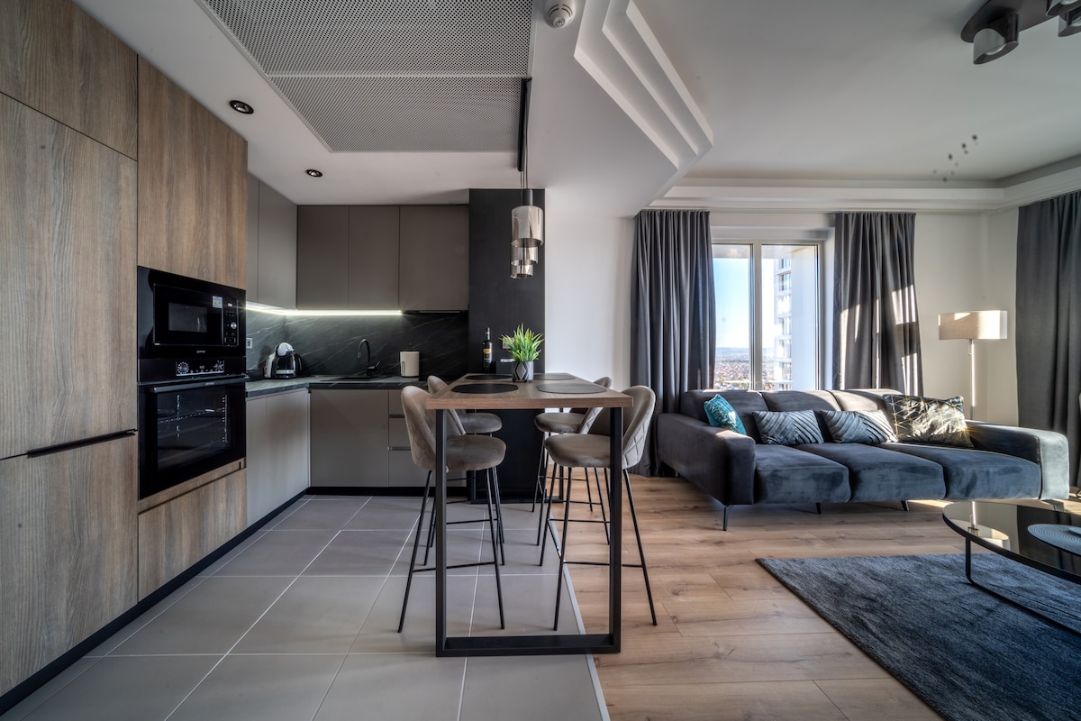A modern kitchen and living area are showcased, featuring wood cabinetry and a dark countertop. A dining table with four high stools is positioned near a large window, allowing natural light to illuminate the space. A comfortable gray sofa and a coffee table complete the inviting setting.
