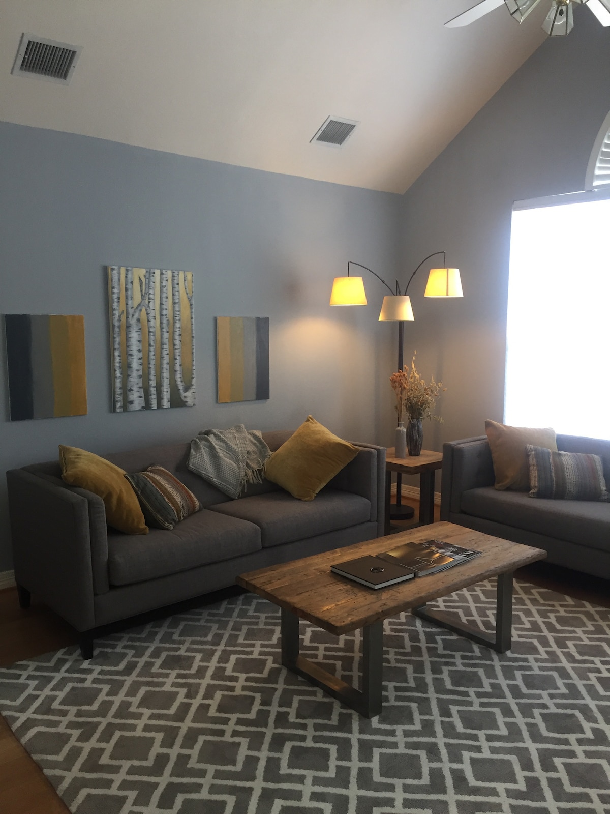 A welcoming living area features two gray sofas adorned with patterned and solid throw pillows. A wooden coffee table sits between them, complemented by decorative items. The space is illuminated by two arc floor lamps, and natural light enters through a large window covered with sheer shades.