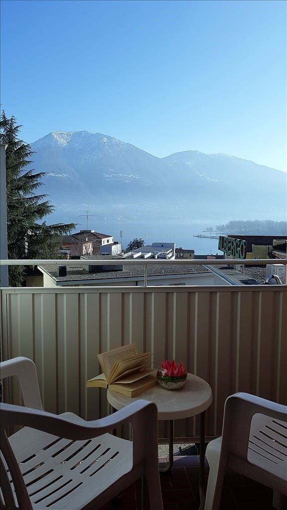 A small balcony is visible, featuring two white chairs and a round table. A plant and a book rest on the table. In the background, mountains rise against a clear blue sky, with the lake glistening below.