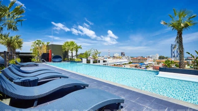 A rooftop pool area is visible, featuring a long infinity pool surrounded by palm trees and lounge chairs. The clear blue sky and city skyline can be seen in the background, creating a serene atmosphere for relaxation.