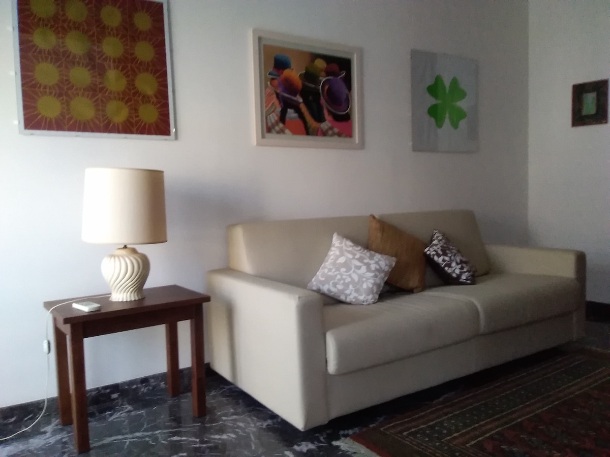 A light-colored sofa is featured with decorative pillows, positioned beside a small wooden table. A table lamp stands on the table, providing soft illumination. Colorful artworks adorn the walls, adding visual interest to the space.