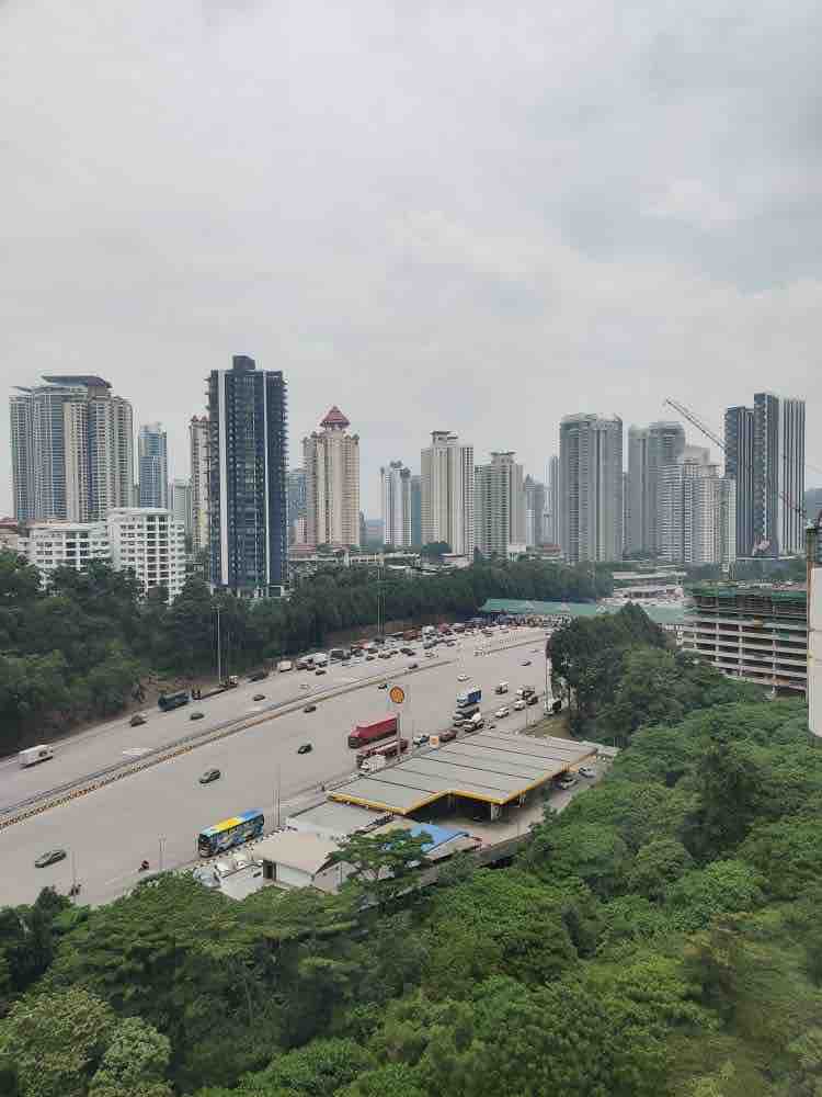 A city view captured from a height shows a busy road lined with vehicles, surrounded by lush greenery. Skyscrapers dominate the skyline, showcasing a range of architectural styles under a cloudy sky.