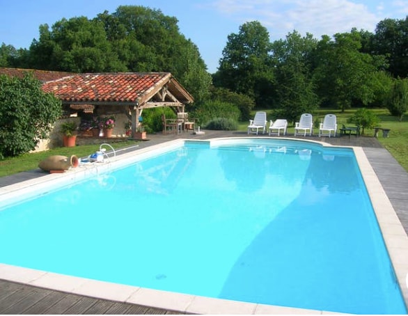 A refreshing swimming pool is surrounded by a wooden deck, with sun loungers positioned for relaxation. Lush greenery and trees form a serene backdrop, while a rustic structure with a tiled roof offers shaded seating nearby.