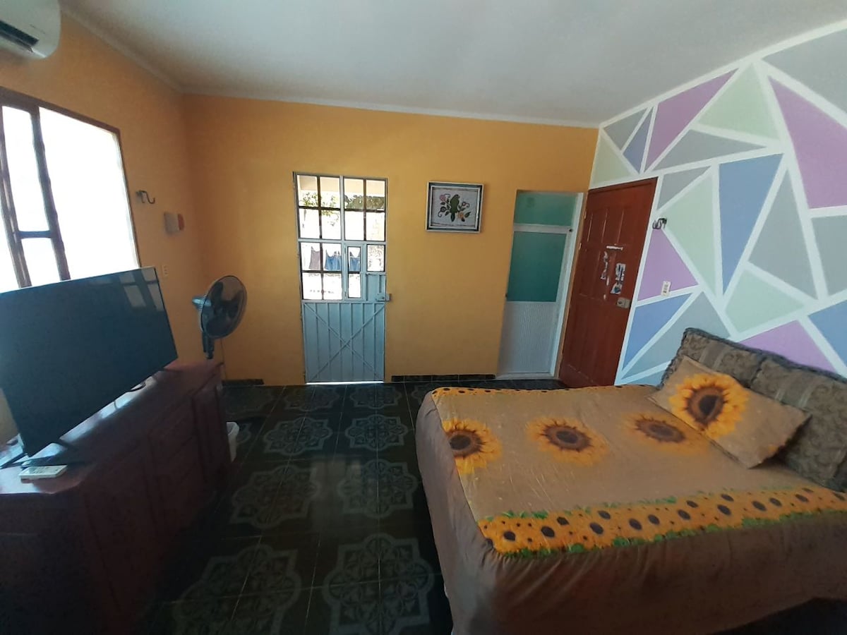 A bright room features orange walls and colorful geometric accents. A spacious bed with a sunflower design is positioned near a window, allowing natural light. A small television sits on a wooden cabinet, while a fan is positioned beside the bed for comfort.