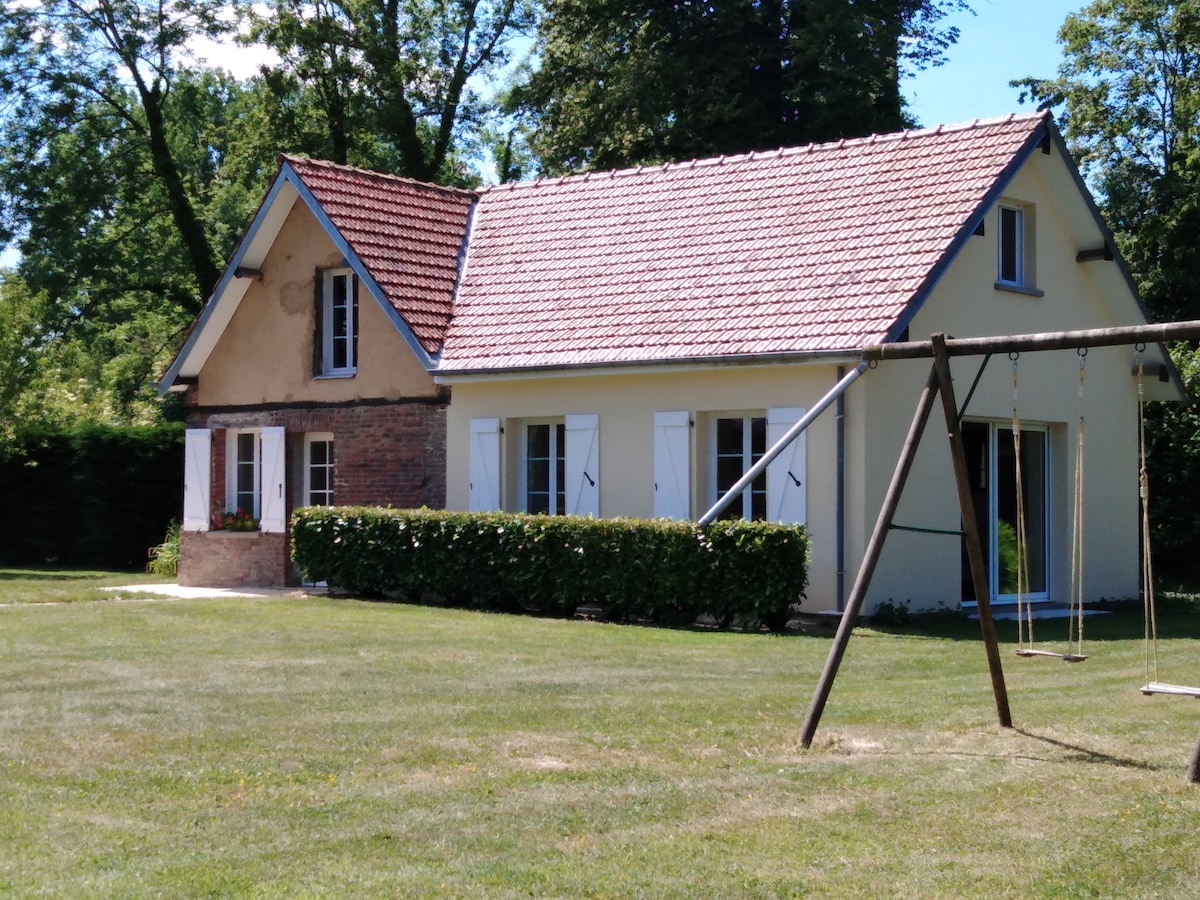 The exterior of a charming house is surrounded by greenery, featuring a sloped roof and a mix of brick and light-colored siding. A swing set stands on the lawn, providing a playful element to the spacious garden area.