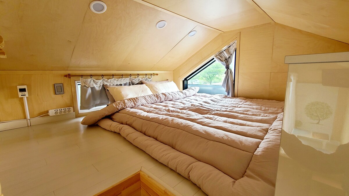 A lofted sleeping area is featured with a spacious bed dressed in soft, neutral-toned bedding. Natural light filters in through a window with stylish drapery, highlighting the warm wooden tones of the room.
