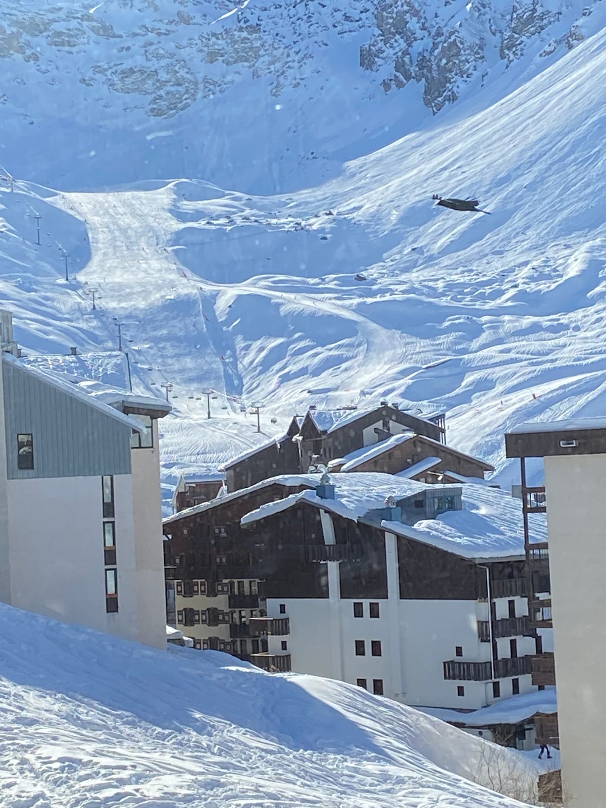 Snow-covered ski slopes are visible in the background, with a variety of snow-laden buildings in the foreground. A clear sky above enhances the crisp winter scene, while the mountain landscape provides a scenic backdrop for the area.