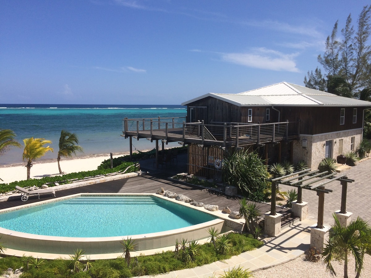The image captures a seaside property featuring a wooden deck overlooking the ocean. A sunlit swimming pool is situated in front, framed by lush greenery. The clear blue waters stretch into the horizon, with sandy beach areas visible on either side of the deck.