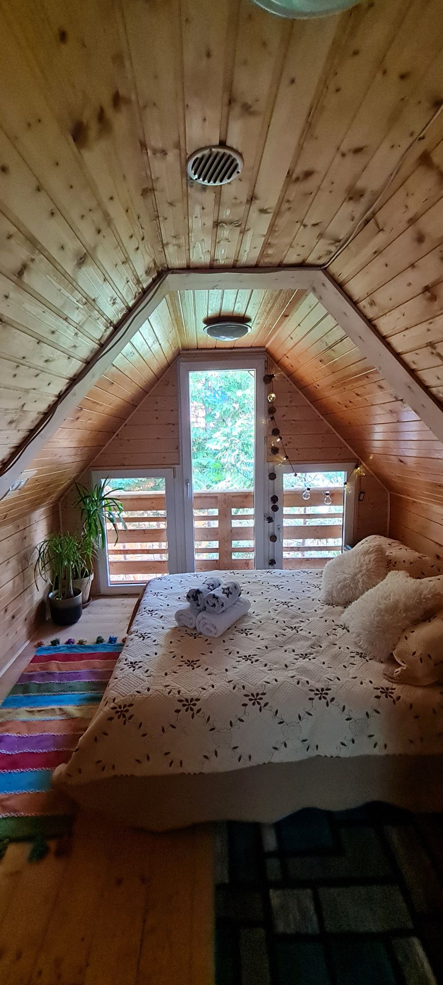 The bedroom features a sloped wooden ceiling complemented by large windows that provide natural light and views of greenery. A comfortable bed is adorned with decorative pillows and a light blanket. A colorful rug adds warmth to the wooden floor, and a potted plant is positioned nearby.