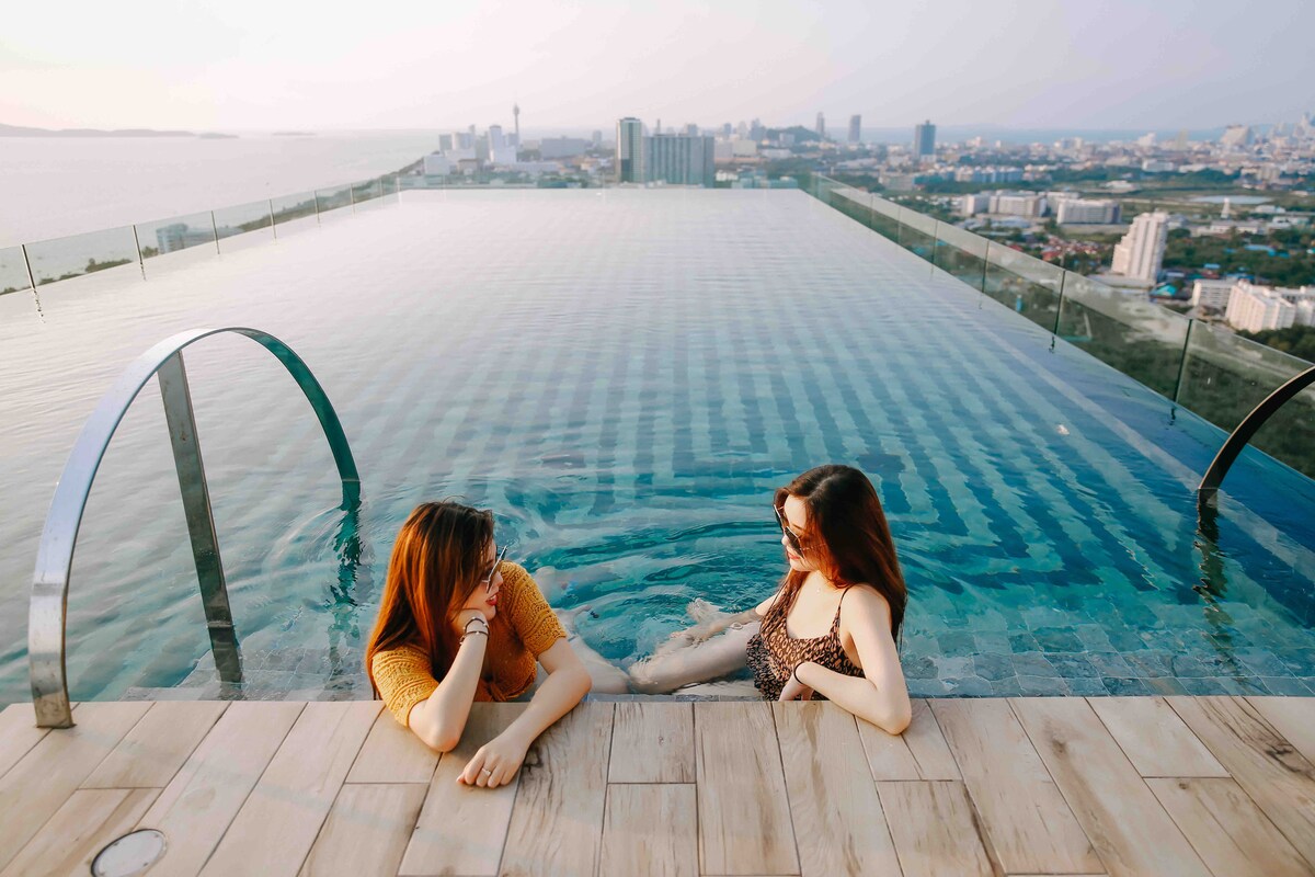 An infinity pool is visible, extending towards the horizon with city views in the background. The smooth wooden deck complements the clear water, while two individuals are seen sitting peacefully at the water's edge, relaxing and enjoying the atmosphere.