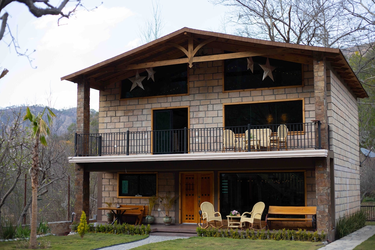 The two-story house features a stone exterior with wooden accents and a spacious balcony. A comfortable seating area is visible on the balcony, while a well-maintained lawn leads to the entrance. Surrounding greenery and distant mountains create a serene setting.