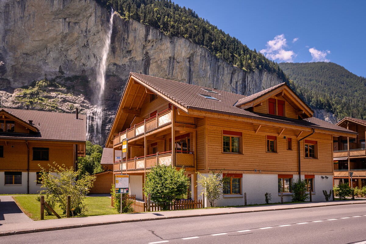 The apartment building features a modern wooden exterior with balconies, set against a backdrop of a towering mountain and cascading waterfall. Lush greenery and signage are noted in the foreground, highlighting the inviting atmosphere of the Lauterbrunnen valley.