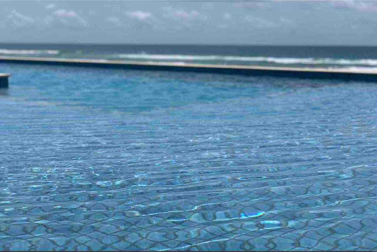 An expansive infinity pool features crystal-clear water reflecting the sky, with gentle waves of the ocean visible in the background. The pool edges blend into the horizon, creating a seamless view of the landscape.