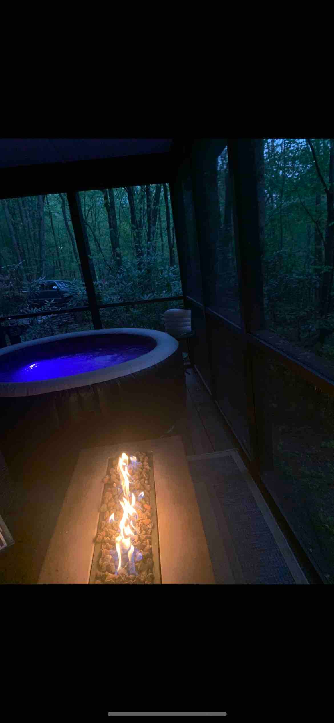 An inflatable hot tub sits on a screened-in porch, illuminated in soft blue light. Nearby, a gas fire table features flickering flames, with surrounding views of lush greenery visible through the screened walls, creating a relaxing atmosphere for evening gatherings.