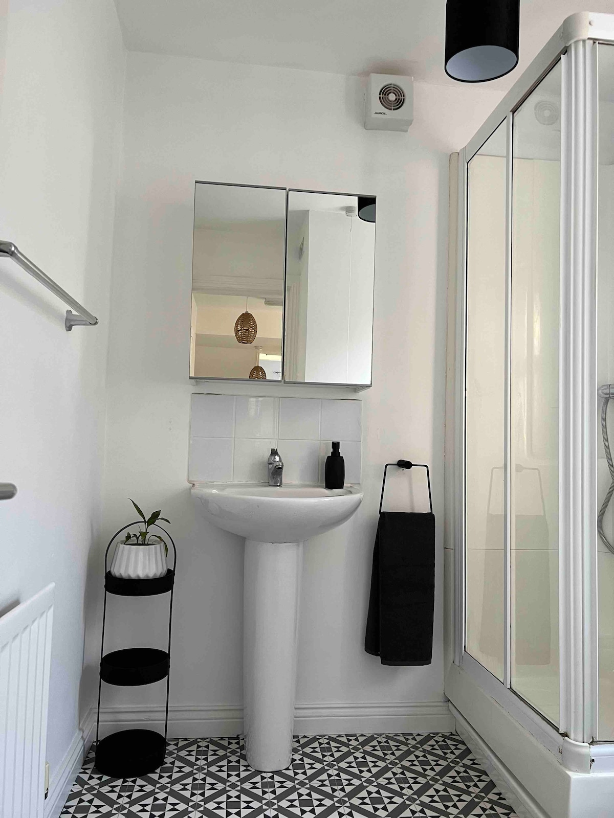 A modern bathroom features a compact sink with a mirror above it. A shower enclosure is visible on one side, while a stylish black and white tiled floor adds visual interest. A small plant sits on a stand for a touch of greenery.
