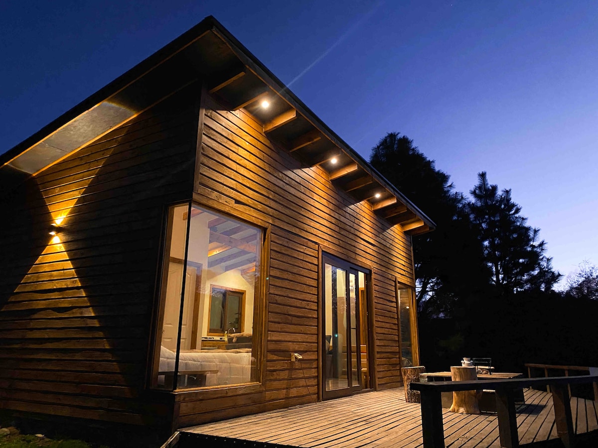The exterior of a tiny house is shown at dusk, featuring wooden siding and multiple large windows. Soft lighting highlights the structure's angles and inviting deck area, which includes natural wood tables. The surrounding greenery is softly illuminated by the evening light.