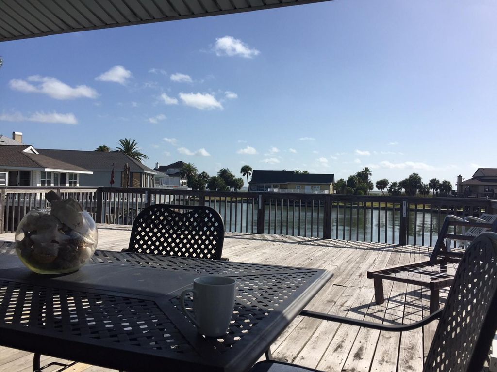 A spacious deck overlooks a serene canal, with a table and chairs providing seating for relaxation. A large glass bowl filled with shells is placed on the table, while the sun shines brightly against a clear blue sky, enhancing the tranquil water view.