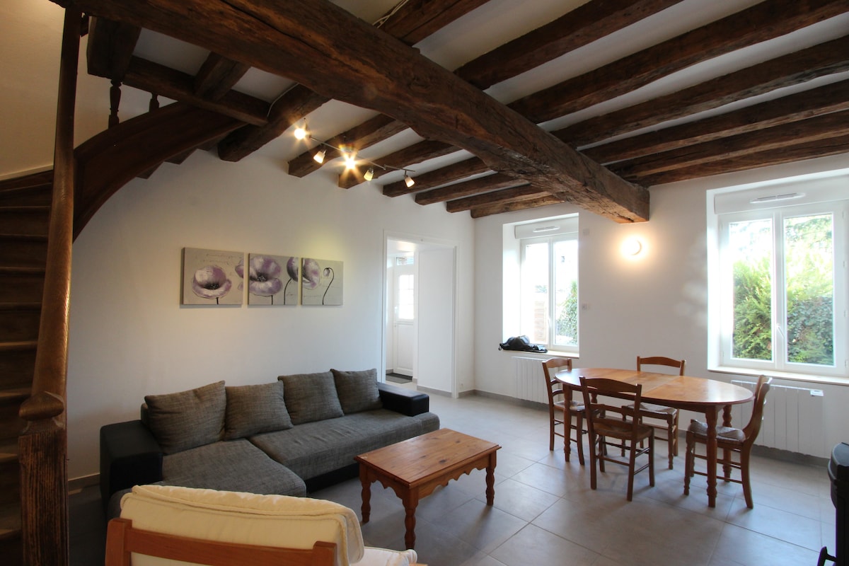 An open living area features exposed wooden beams on the ceiling. A sectional sofa is positioned beside a wooden coffee table. A round dining table with wooden chairs sits near large windows, allowing natural light to illuminate the space.