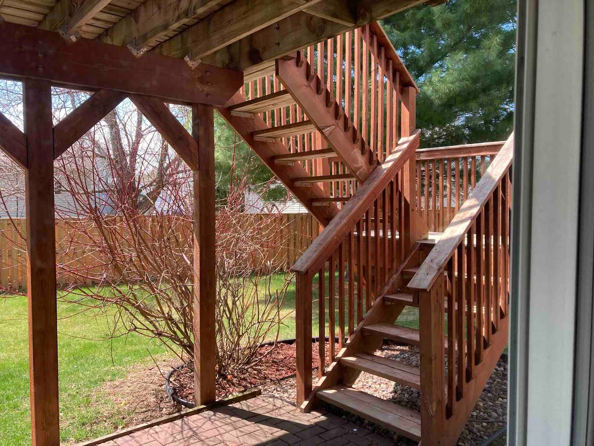 A wooden staircase is seen ascending alongside a well-maintained yard, surrounded by a wooden fence. Vibrant green grass covers the ground, with a small bush visible nearby. Sunlight filters through the trees, casting gentle shadows on the steps.