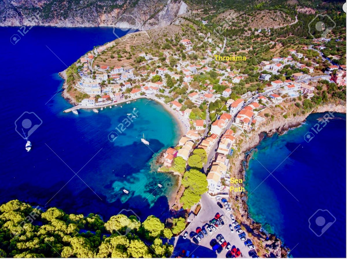 An aerial view captures the coastal village of Asos, showcasing a blend of colorful buildings and lush greenery. The clear blue waters are visible, with several boats anchored. The surrounding hills provide a natural backdrop, highlighting the tranquil environment of the area.