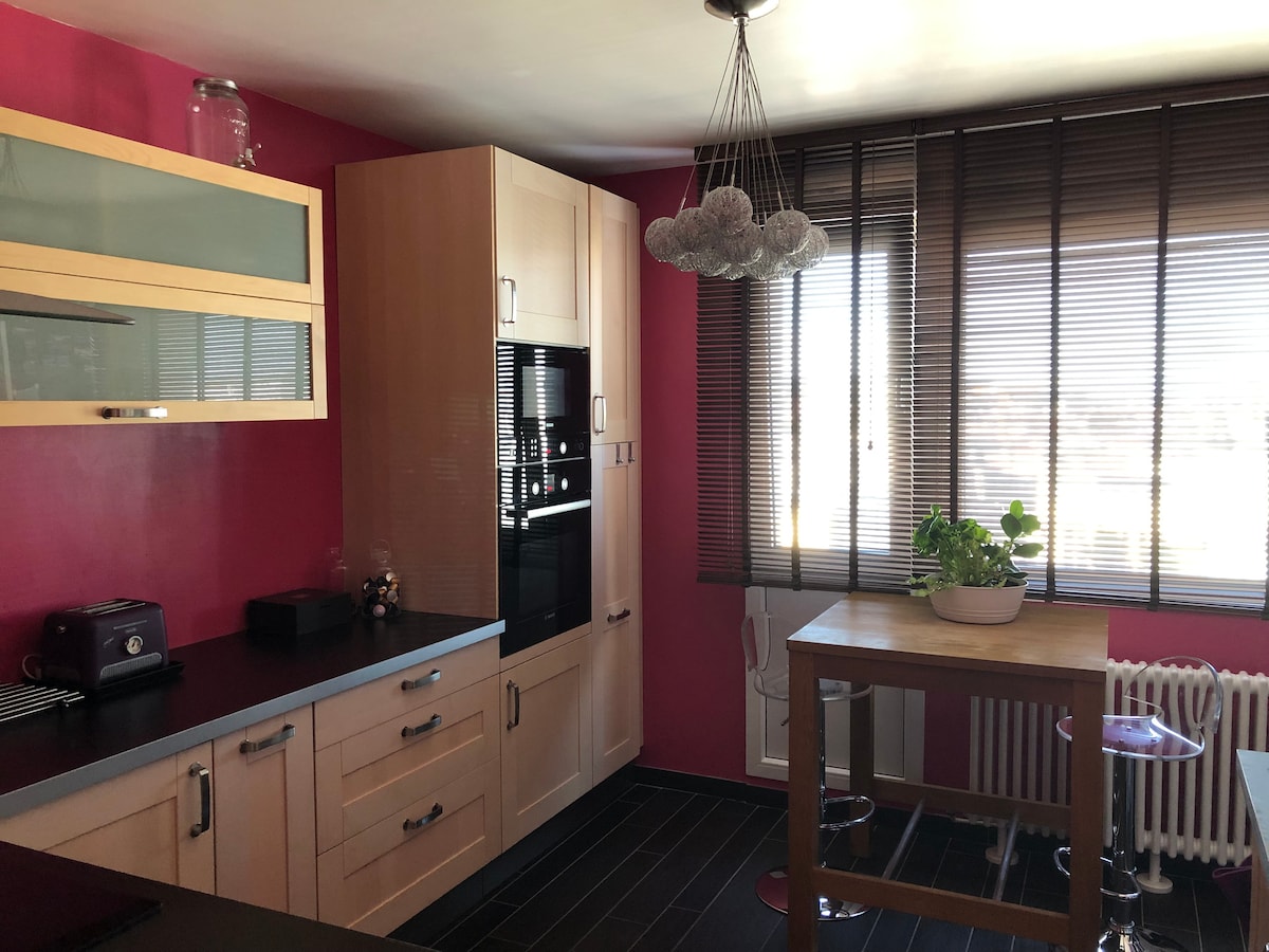 The kitchen area features a combination of wooden cabinets and modern appliances, including an integrated oven and a toaster. A round wooden table is nestled beside a window with blinds, allowing natural light to filter in. A small plant adds a touch of greenery to the space.