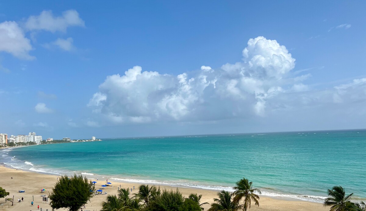 A panoramic view captures a sandy beach bordered by gentle waves and a tranquil blue ocean. Puffy clouds drift across a vibrant sky, while beach chairs and palm trees are visible along the shore, adding to the serene coastal atmosphere.