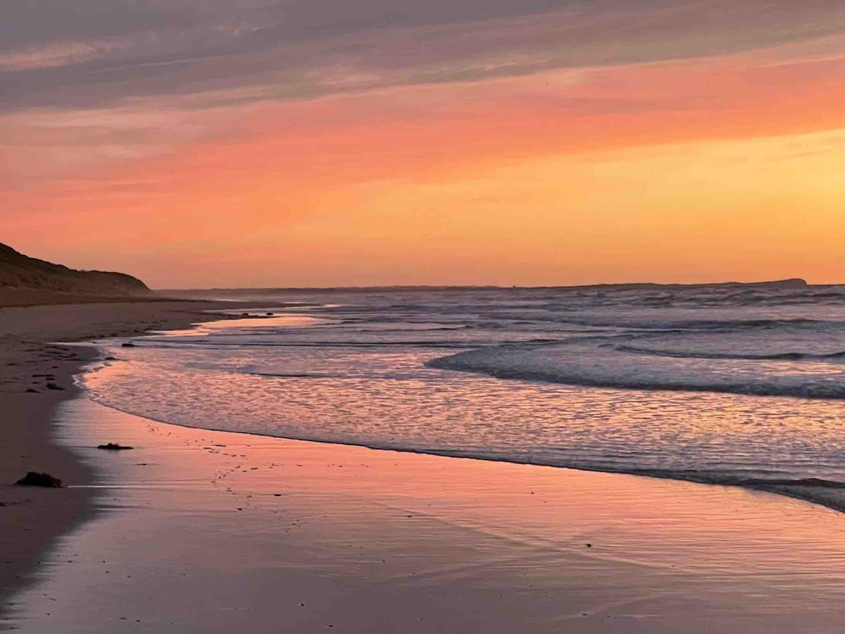 A serene beach scene is captured at sunset, showcasing gentle waves lapping at the shore. The sand reflects soft pink and orange hues from the sky, while distant hills frame the horizon. The tranquil atmosphere invites a sense of peace and relaxation.