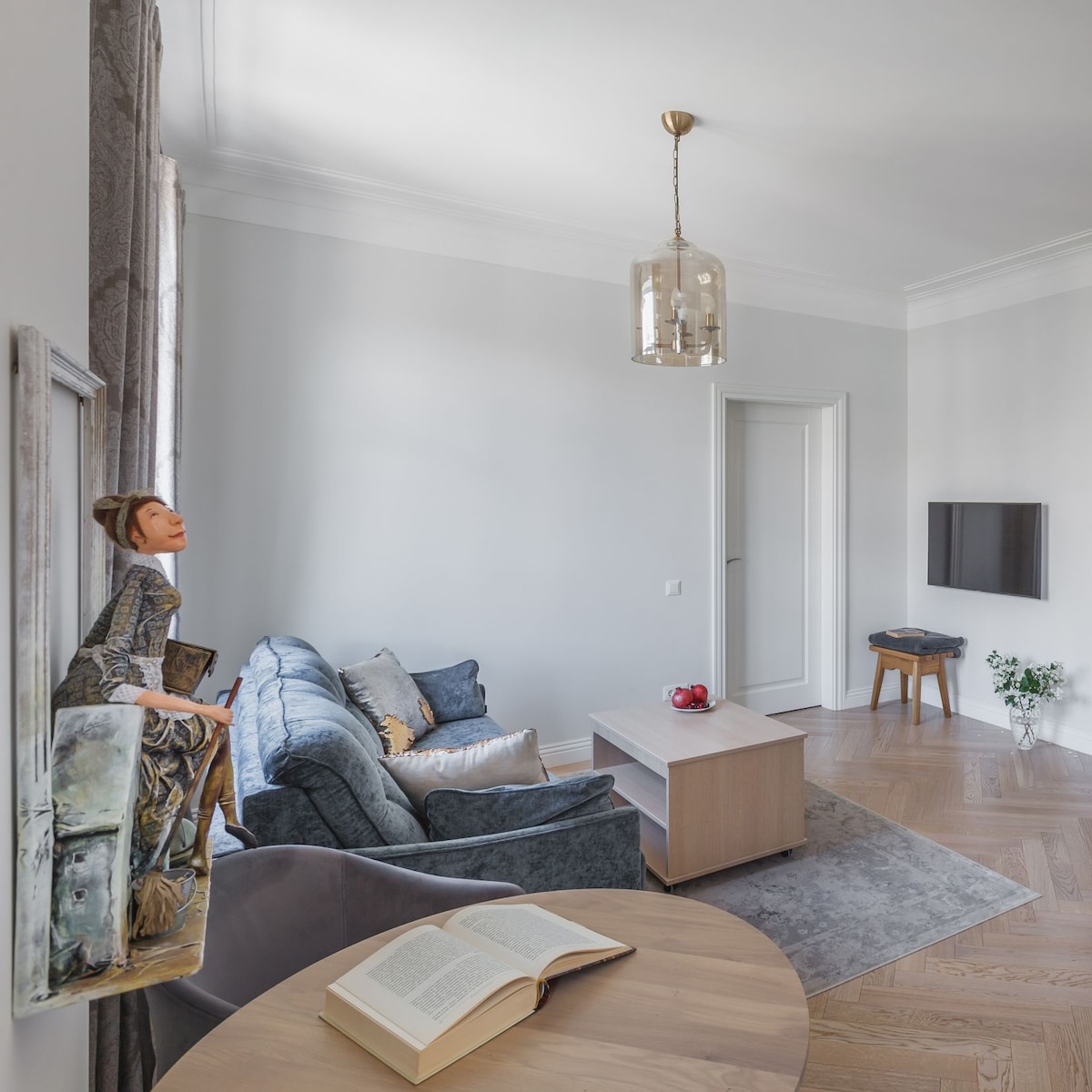 A well-lit living area features a comfortable sofa with cushions. A small coffee table is centrally placed on a patterned rug. Light streams in through large windows, highlighting the soft color palette of the walls and furnishings.
