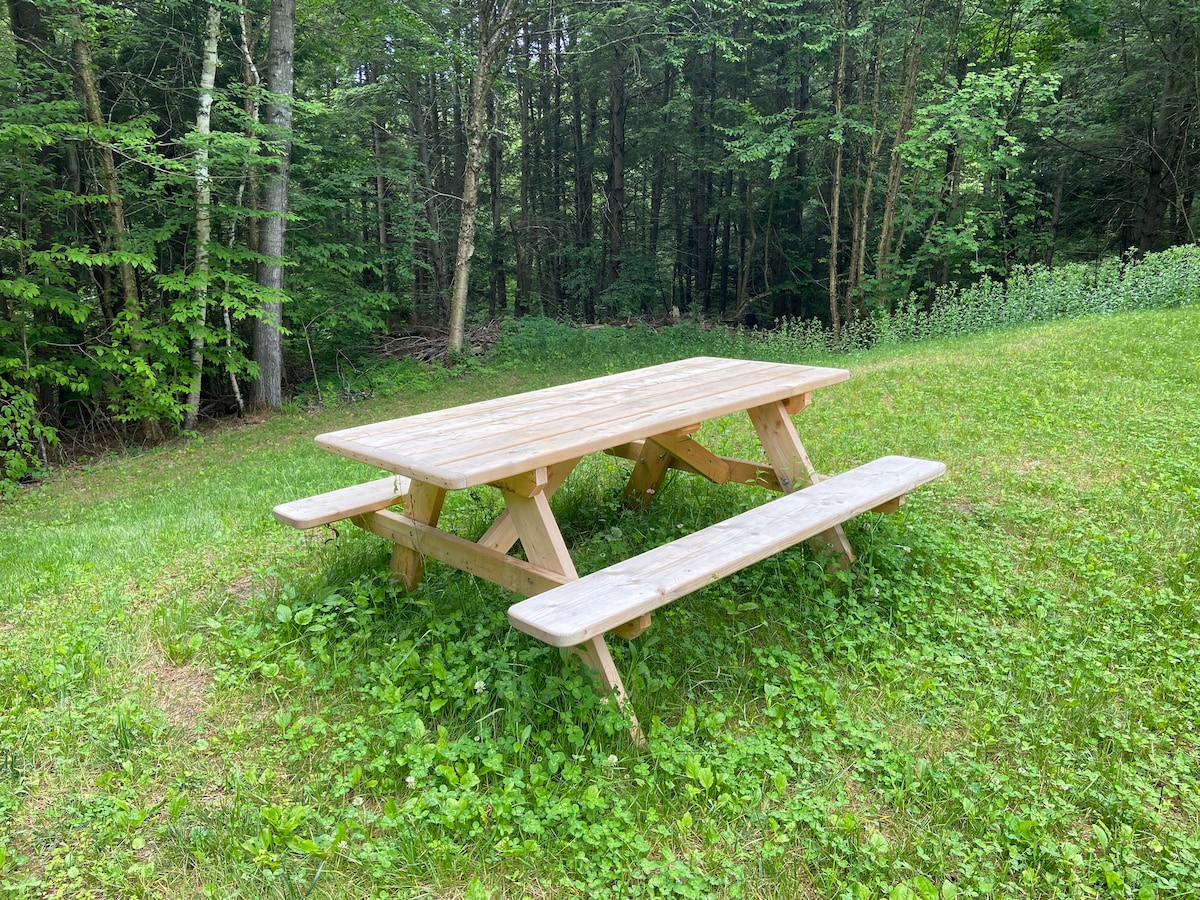 A wooden picnic table is situated on a grassy lawn, surrounded by lush greenery and trees. Two benches are positioned on either side of the table, providing ample seating for outdoor meals or gatherings.
