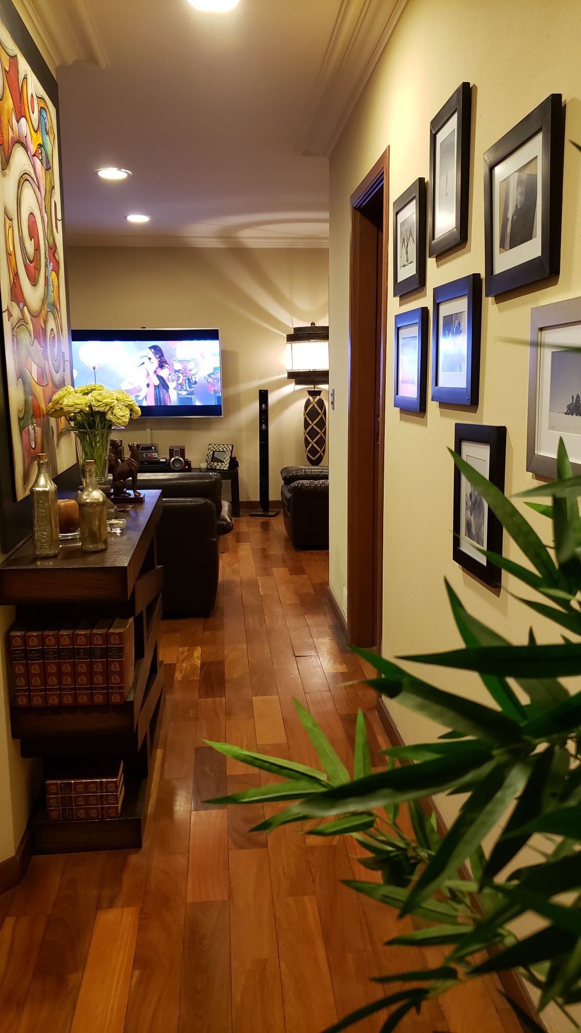 A hallway features polished wood flooring, with a vibrant artwork displayed prominently on the wall. Framed pictures are arranged along the opposite wall. A cozy sitting area can be seen, with a television and warm lighting creating an inviting ambiance.