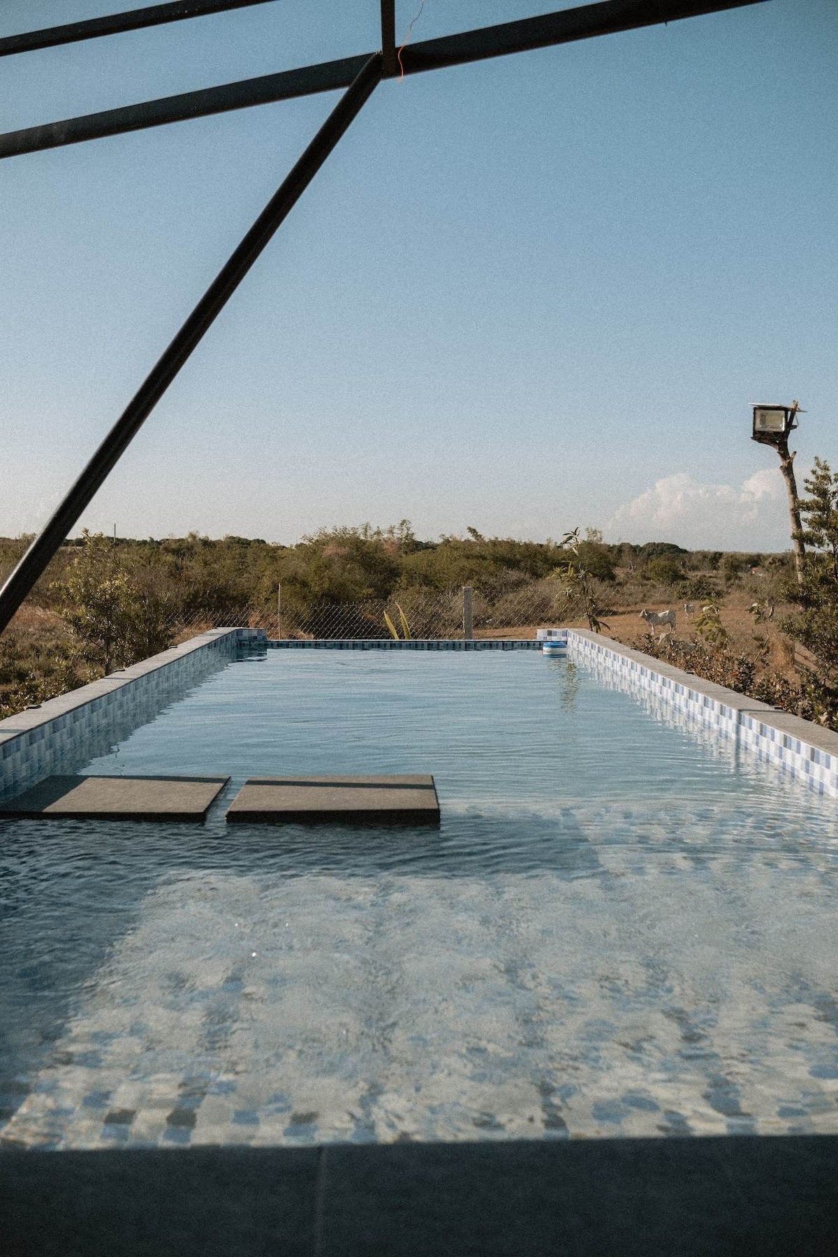 An outdoor pool features clear water reflecting the sky, surrounded by natural scenery. Two stone loungers are positioned at the pool's edge, creating a relaxing area for guests. A fence provides privacy, while a distant light post is visible against the horizon.