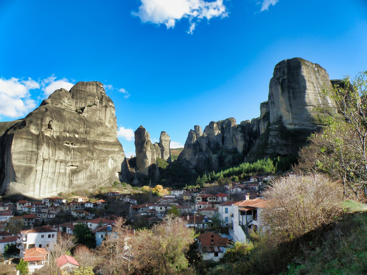 A panoramic view captures towering rock formations against a clear blue sky, with a village nestled at their base. The landscape showcases a mix of rugged cliffs and greens, highlighting the natural beauty surrounding the area.
