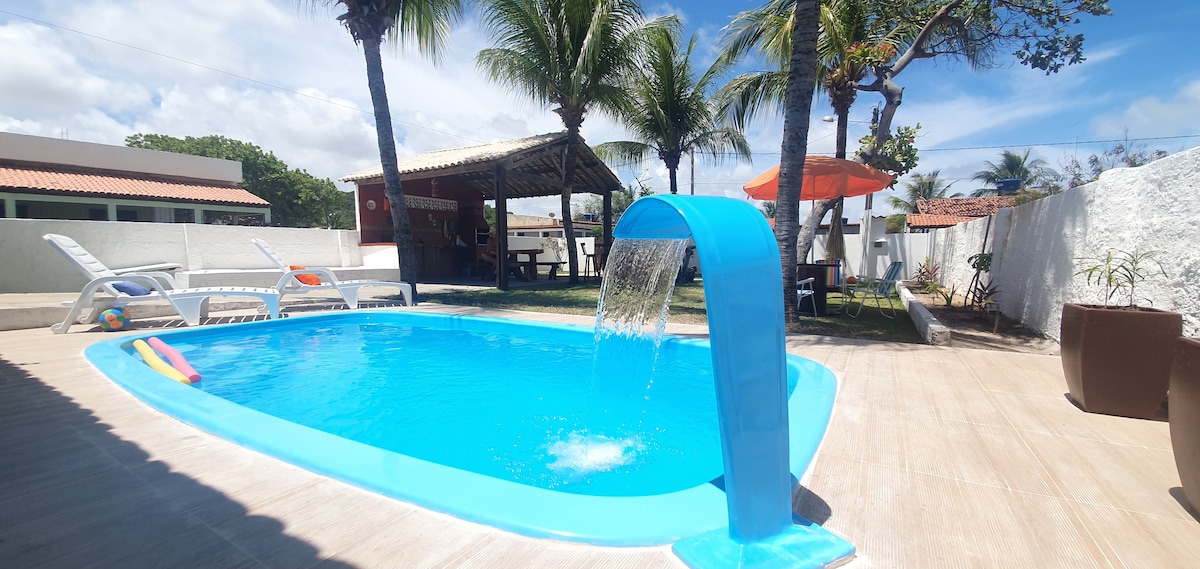 A swimming pool with a water feature flows gently into a vibrant blue. Surrounding greenery includes palm trees and a shaded area with a thatched roof. Lounge chairs are positioned nearby, offering space for relaxation.