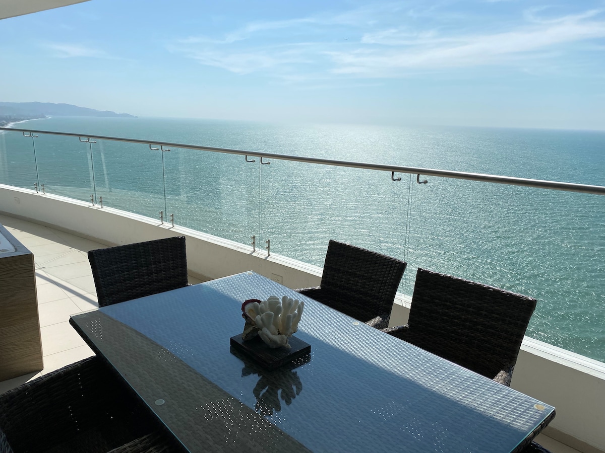 A spacious terrace is shown, featuring a glass tabletop surrounded by four woven chairs. The backdrop reveals a panoramic ocean view, with sunlight shimmering on the water, and a decorative centerpiece on the table adds a subtle touch.