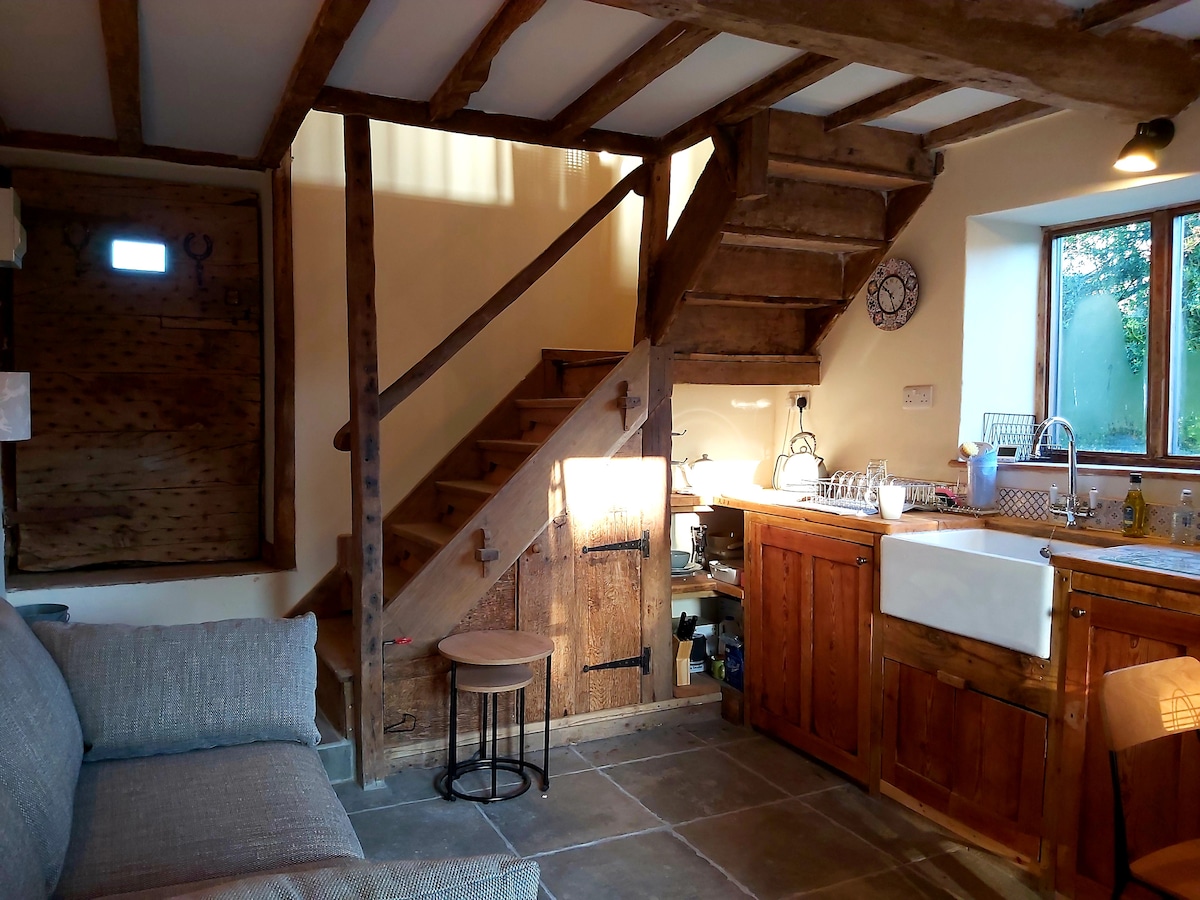 An inviting space features rustic wooden beams and a staircase leading to the upper level. A cozy seating area includes a soft gray sofa. The well-equipped kitchen is highlighted by a large farmhouse sink and warm wood cabinetry, with natural light streaming through a window.
