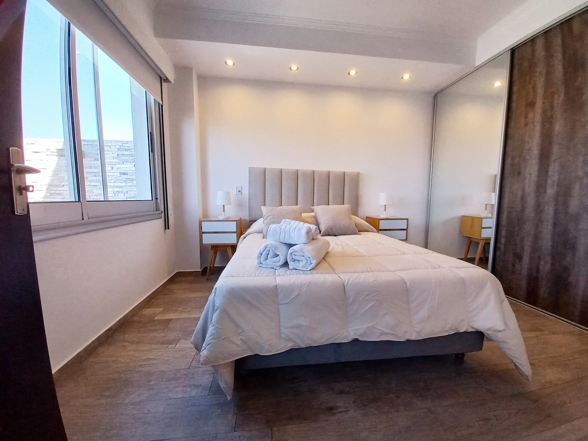 A well-lit bedroom features a spacious bed with a neatly arranged white comforter and two plush pillows. Nightstands with lamps are positioned on either side of the bed. Large windows allow natural light to fill the space, and a mirrored closet adds depth to the room.