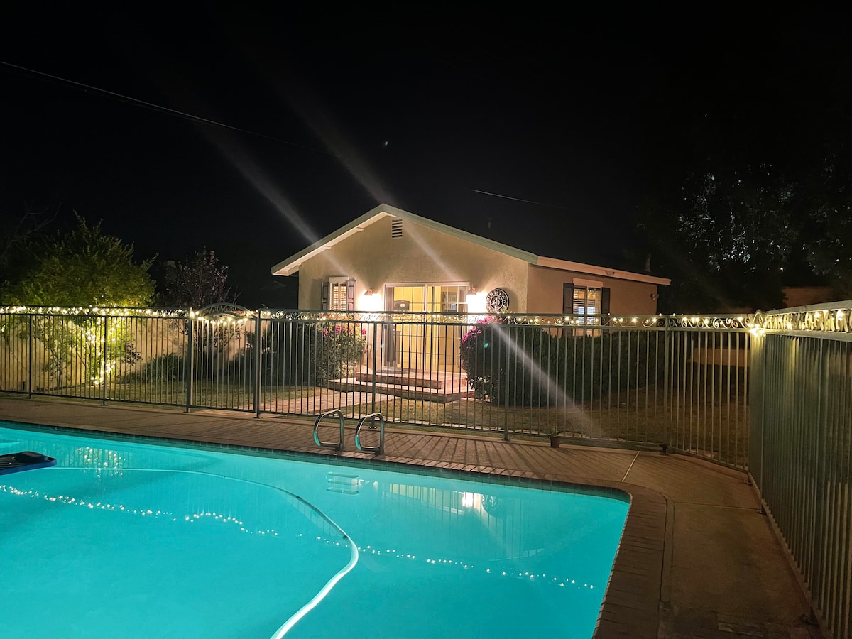 A softly illuminated backyard is highlighted by a gated pool, featuring turquoise water at night. The back house can be seen in the background, surrounded by greenery and soft lights adorning the perimeter. The tranquil setting suggests a peaceful atmosphere.